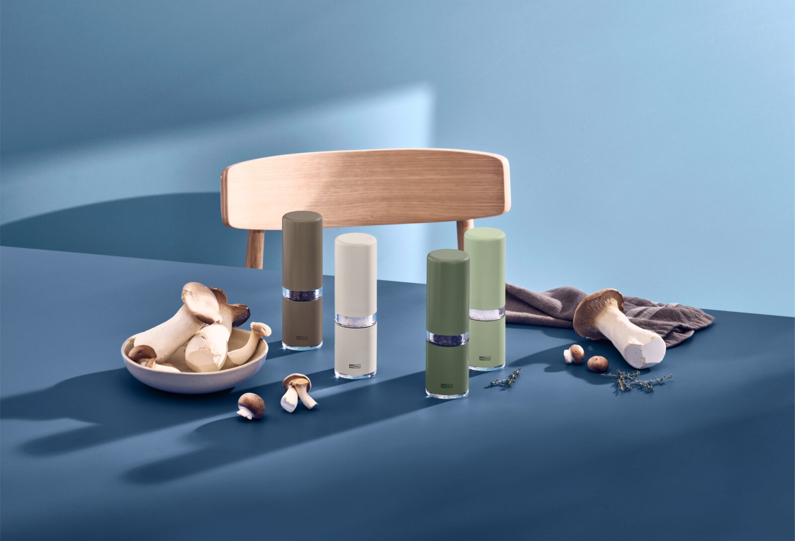 Vier elektrische Salz- und Pfeffermühlen in Grün-, Grau- und Weißtönen auf blauem Tisch, mit Pilzen und Kräutern dekoriert; Perspektive: frontale leicht erhöhte Ansicht.