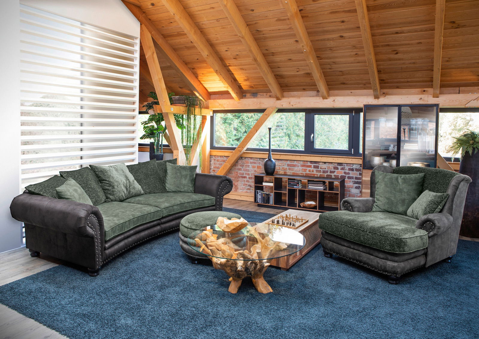 Großes, rundes Megasofa in einem modernen Wohnzimmer mit Holzbalkendecke, grünen Kissen und einem Couchtisch aus Glas und Holz. Perspektive von vorne.