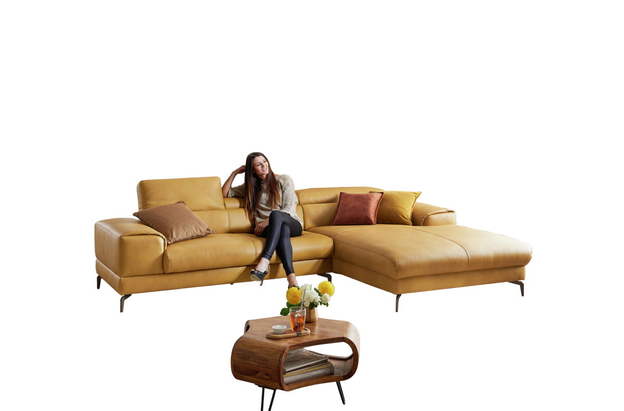 Gelbes Longchair-Sofa mit Kissen, auf dem eine Frau sitzt, aus frontaler Perspektive. Im Vordergrund ein kleiner Holztisch mit Blumen.