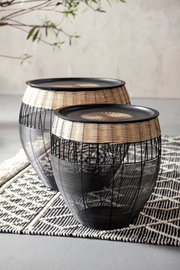 Zwei runde Beistelltische aus schwarzem Metall und hellem Rattan, auf einem gemusterten Teppich stehend, seitliche Perspektive.