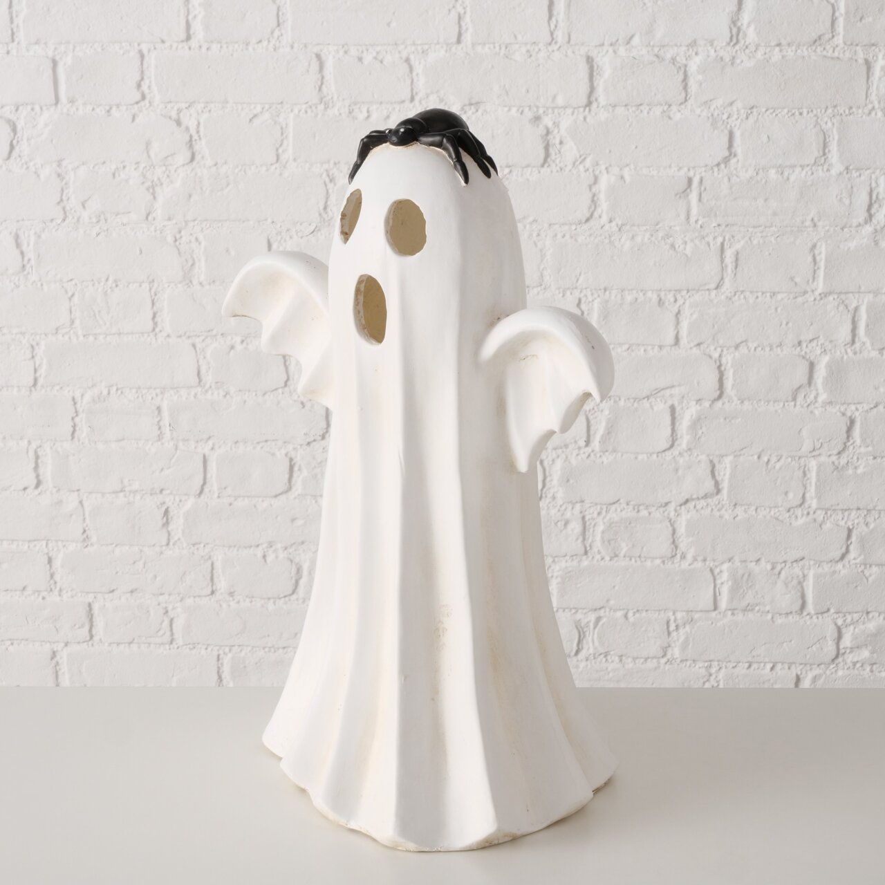 Weiße Halloween-Geisterfigur mit ausgebreiteten Armen und schwarzer Spinne auf dem Kopf vor weißer Ziegelwand, frontal in leichter Untersicht fotografiert