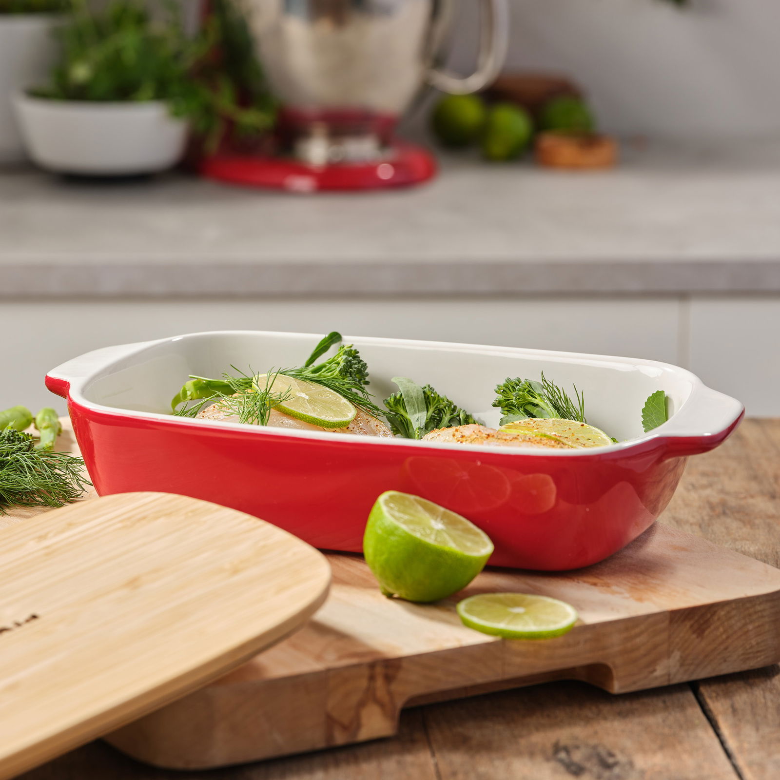 Rote Auflaufform mit Deckel, gefüllt mit frischen Zutaten, auf einem Holzbrett in einer Küche, seitliche Perspektive