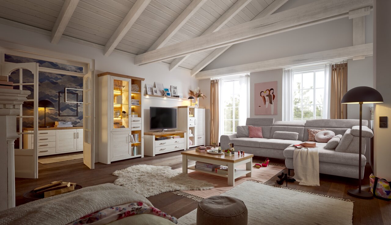 Wohnzimmer mit gemütlicher Einrichtung, bestehend aus einem großen Ecksofa, einem Couchtisch und einer Wohnwand mit Vitrine und TV-Element. Die Perspektive zeigt den Raum von vorne mit Blick auf die gesamte Einrichtung.
