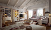 Wohnzimmer mit gemütlicher Einrichtung, bestehend aus einem großen Ecksofa, einem Couchtisch und einer Wohnwand mit Vitrine und TV-Element. Die Perspektive zeigt den Raum von vorne mit Blick auf die gesamte Einrichtung.