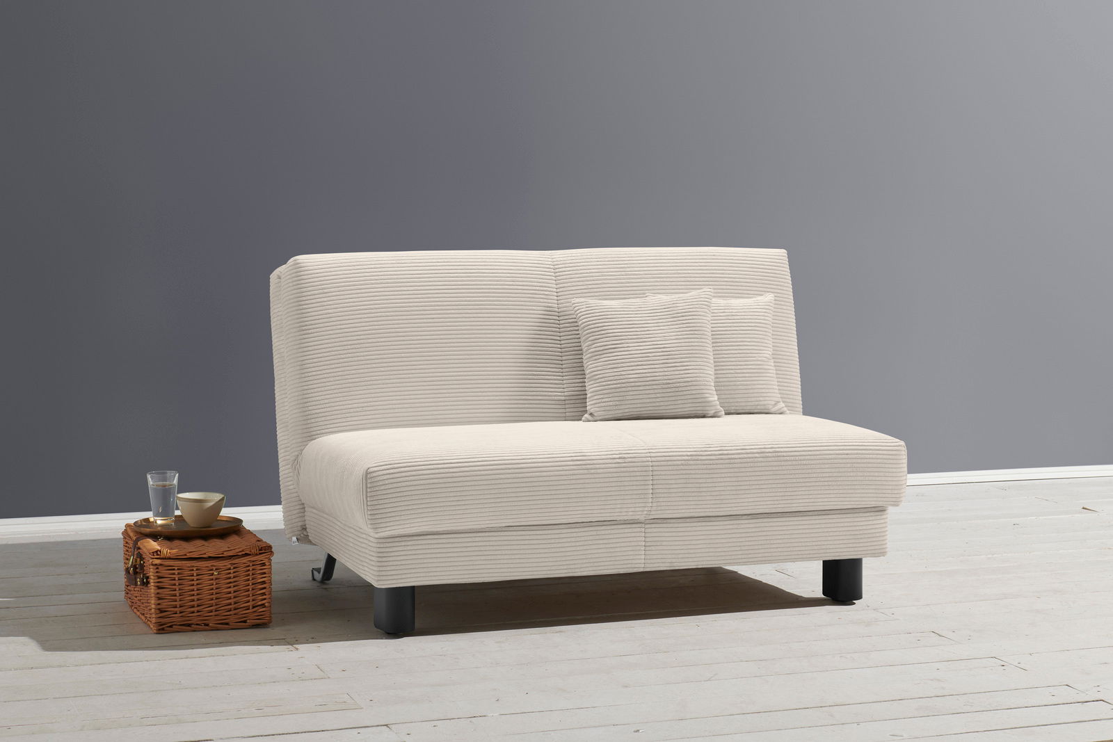 Beiges Funktionssofa mit zwei Kissen, seitliche Perspektive, auf hellem Holzboden vor grauer Wand.