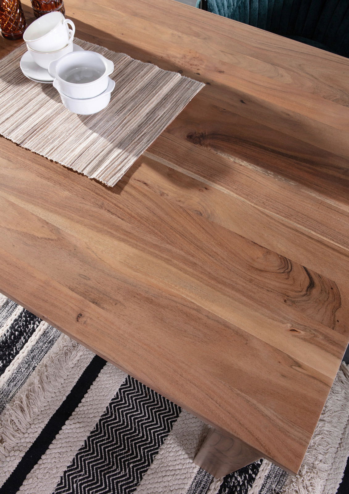 Nahaufnahme eines Esstisches aus Holz mit natürlicher Maserung, darauf stehen weiße Tassen und Schalen auf einem gestreiften Tischläufer, Perspektive von oben.