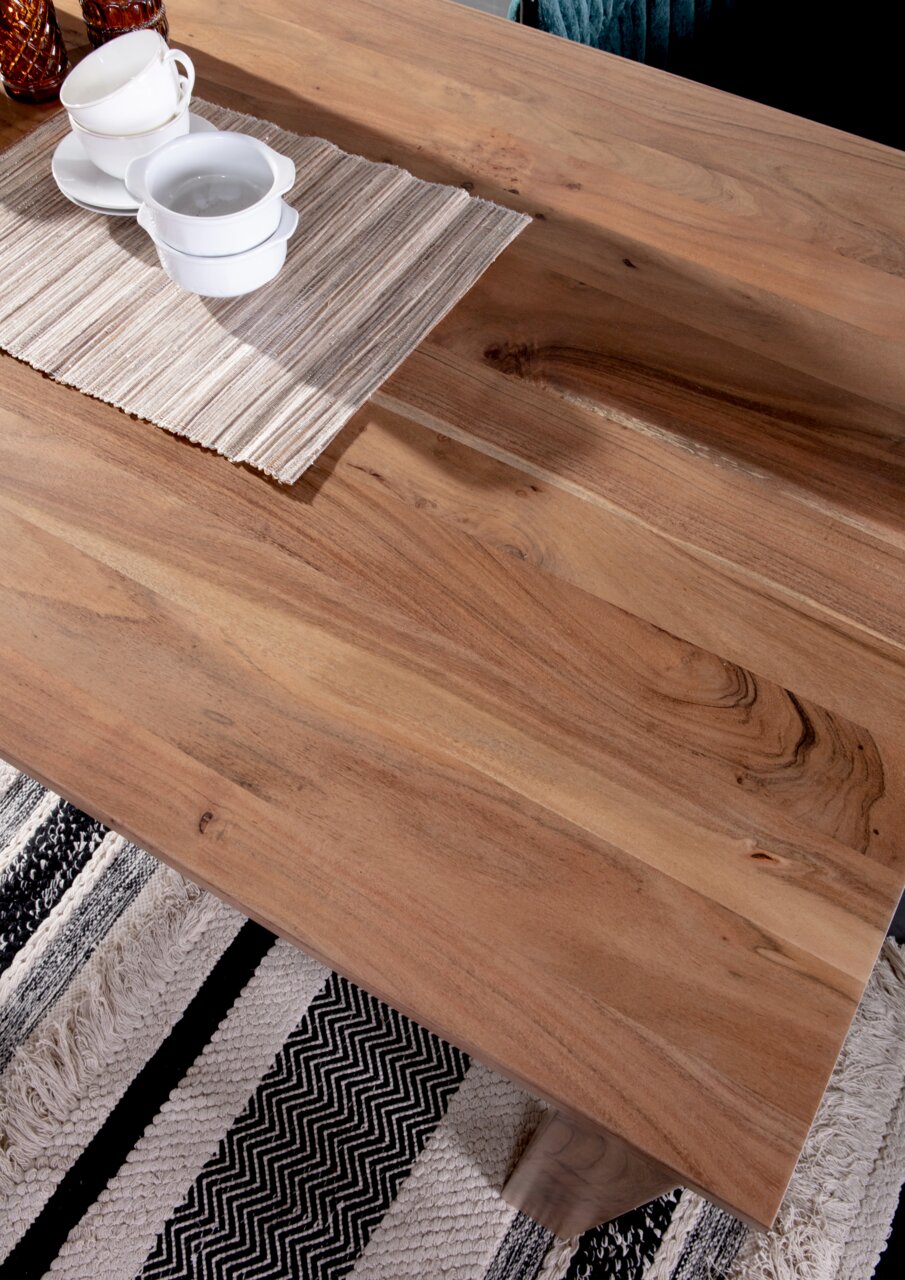 Nahaufnahme eines Esstisches aus Holz mit natürlicher Maserung, darauf stehen weiße Tassen und Schalen auf einem gestreiften Tischläufer, Perspektive von oben.