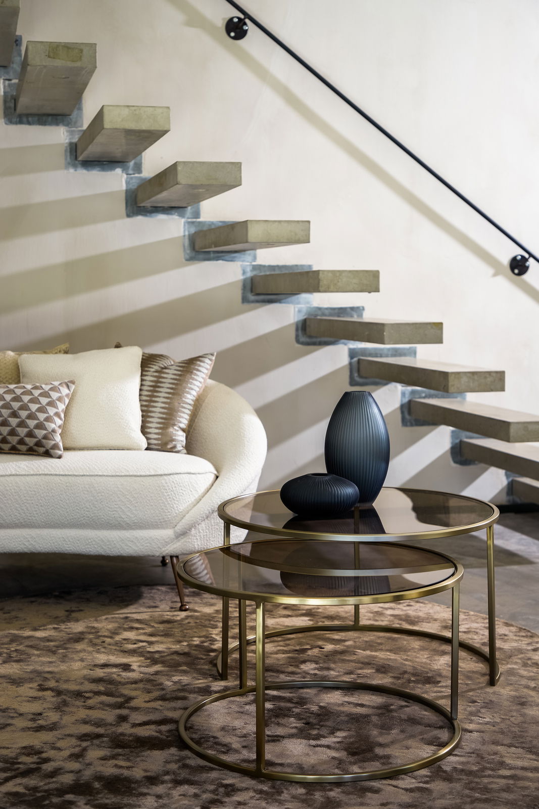Zwei runde Couchtische mit Glasplatten und goldenen Metallrahmen, dekoriert mit zwei dunklen Vasen, vor einer modernen Treppe und einem Sofa, seitliche Perspektive.