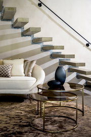 Zwei runde Couchtische mit Glasplatten und goldenen Metallrahmen, dekoriert mit zwei dunklen Vasen, vor einer modernen Treppe und einem Sofa, seitliche Perspektive.