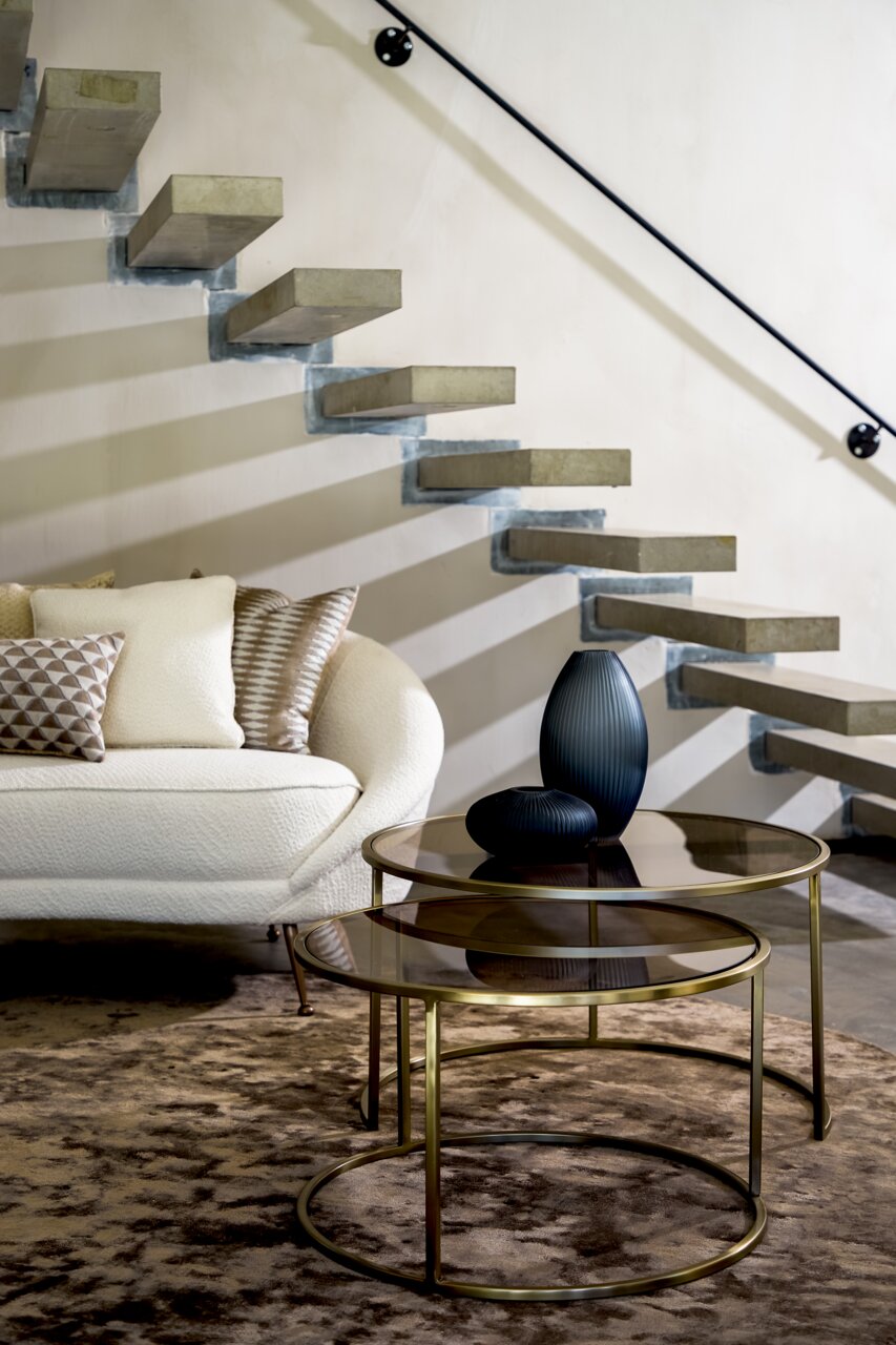 Zwei runde Couchtische mit Glasplatten und goldenen Metallrahmen, dekoriert mit zwei dunklen Vasen, vor einer modernen Treppe und einem Sofa, seitliche Perspektive.