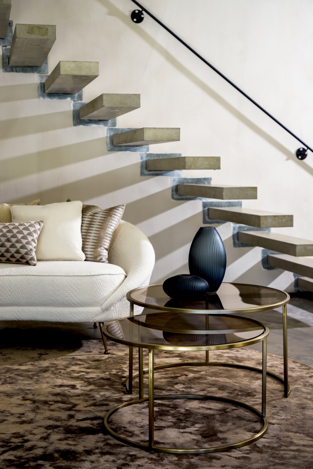 Zwei runde Couchtische mit Glasplatten und goldenen Metallrahmen, dekoriert mit zwei dunklen Vasen, vor einer modernen Treppe und einem Sofa, seitliche Perspektive.