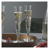 Zwei elegante Champagnergläser mit goldenen und bunten Herzmotiven, gefüllt mit sprudelndem Getränk, auf einem goldenen Tablett. Perspektive: seitlich.