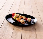 Schwarzer ovaler Teller mit Fleisch- und Gemüsespießen (Tomaten, Paprika, Zwiebeln) auf hellem Holztisch, fotografiert aus leicht erhöhter Schrägansicht.