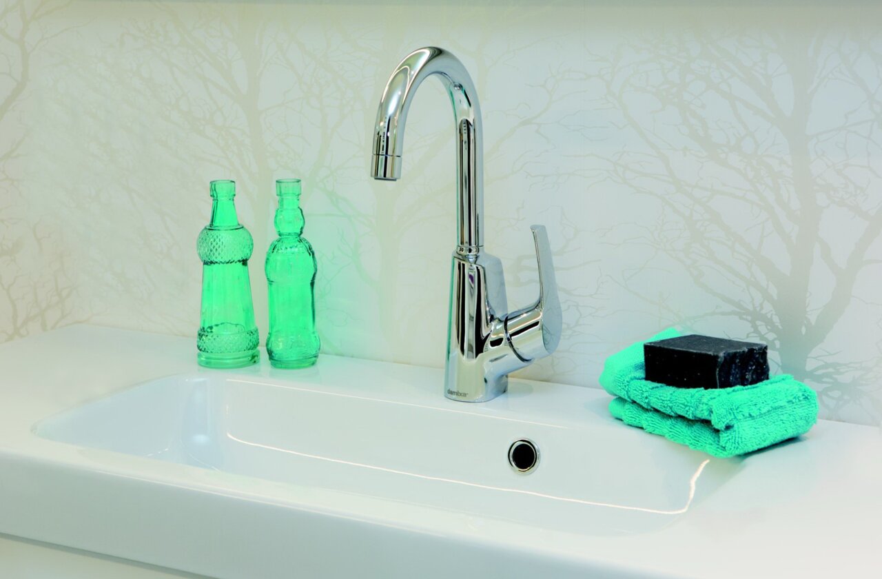 Moderne Waschtischarmatur in Chromoptik, seitliche Perspektive, mit grünen Glasflaschen und Handtuch im Hintergrund.