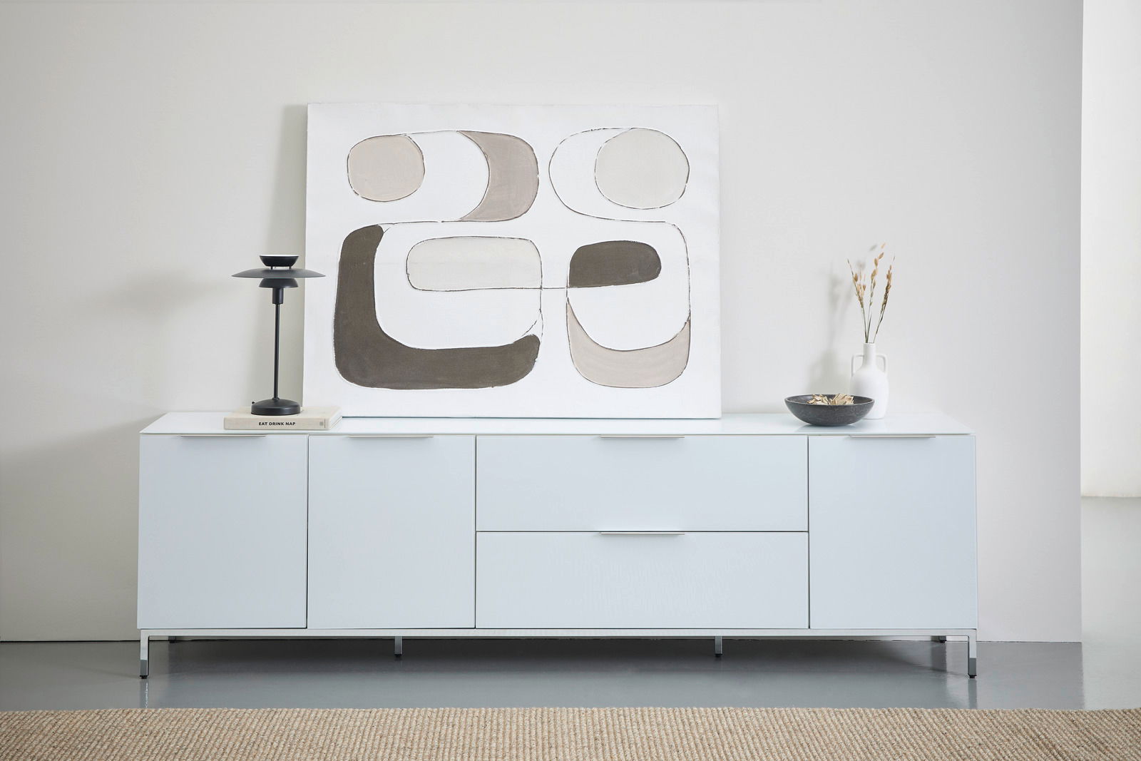 Novel Metalen onderstel 225 MALIN Frontale Ansicht eines modernen, weißen Sideboards mit Metalluntergestell, dekoriert mit einer abstrakten Kunstleinwand, einer schwarzen Lampe, einem Buch, einer Vase und einer Schale.