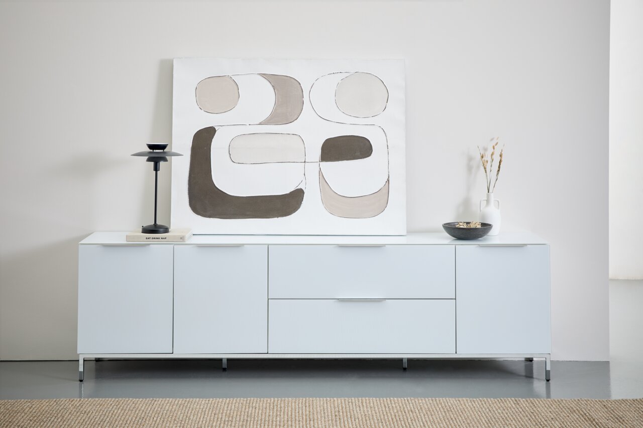 Frontale Ansicht eines modernen, weißen Sideboards mit Metalluntergestell, dekoriert mit einer abstrakten Kunstleinwand, einer schwarzen Lampe, einem Buch, einer Vase und einer Schale.