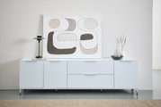 Frontale Ansicht eines modernen, weißen Sideboards mit Metalluntergestell, dekoriert mit einer abstrakten Kunstleinwand, einer schwarzen Lampe, einem Buch, einer Vase und einer Schale.