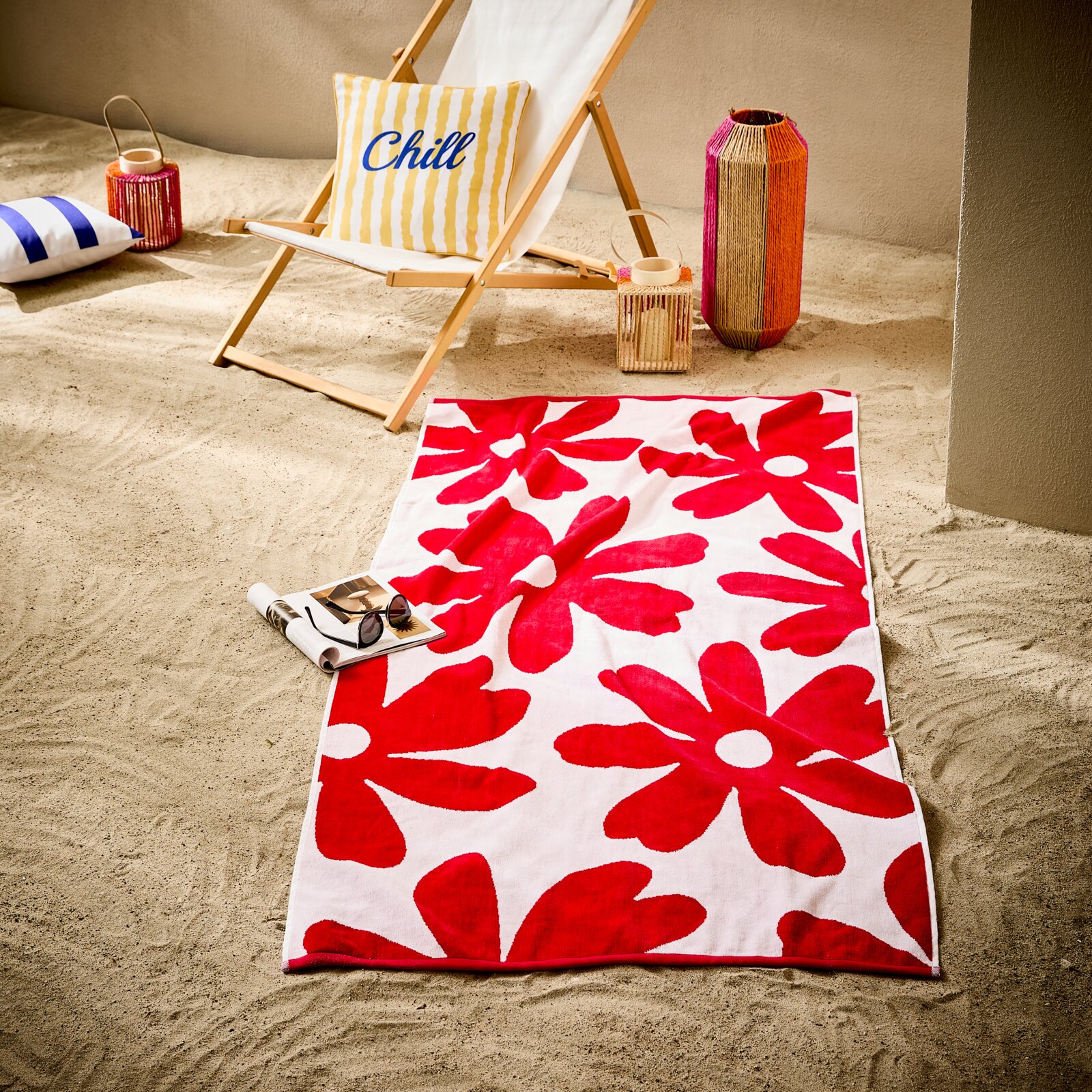 Esposa Strandtuch 90x180cm Rot-weißes Strandtuch mit großem Blumenmuster auf Sand, im Vordergrund mit Zeitschrift und Sonnenbrille; dahinter Liegestuhl mit Kissen sowie dekorative Laternen – Perspektive: leicht von oben, schräg von vorn