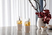 Zwei Longdrink-Gläser mit Getränken und Eiswürfeln auf einem Marmortisch, daneben eine Vase mit lila Orchideen, seitliche Perspektive.
