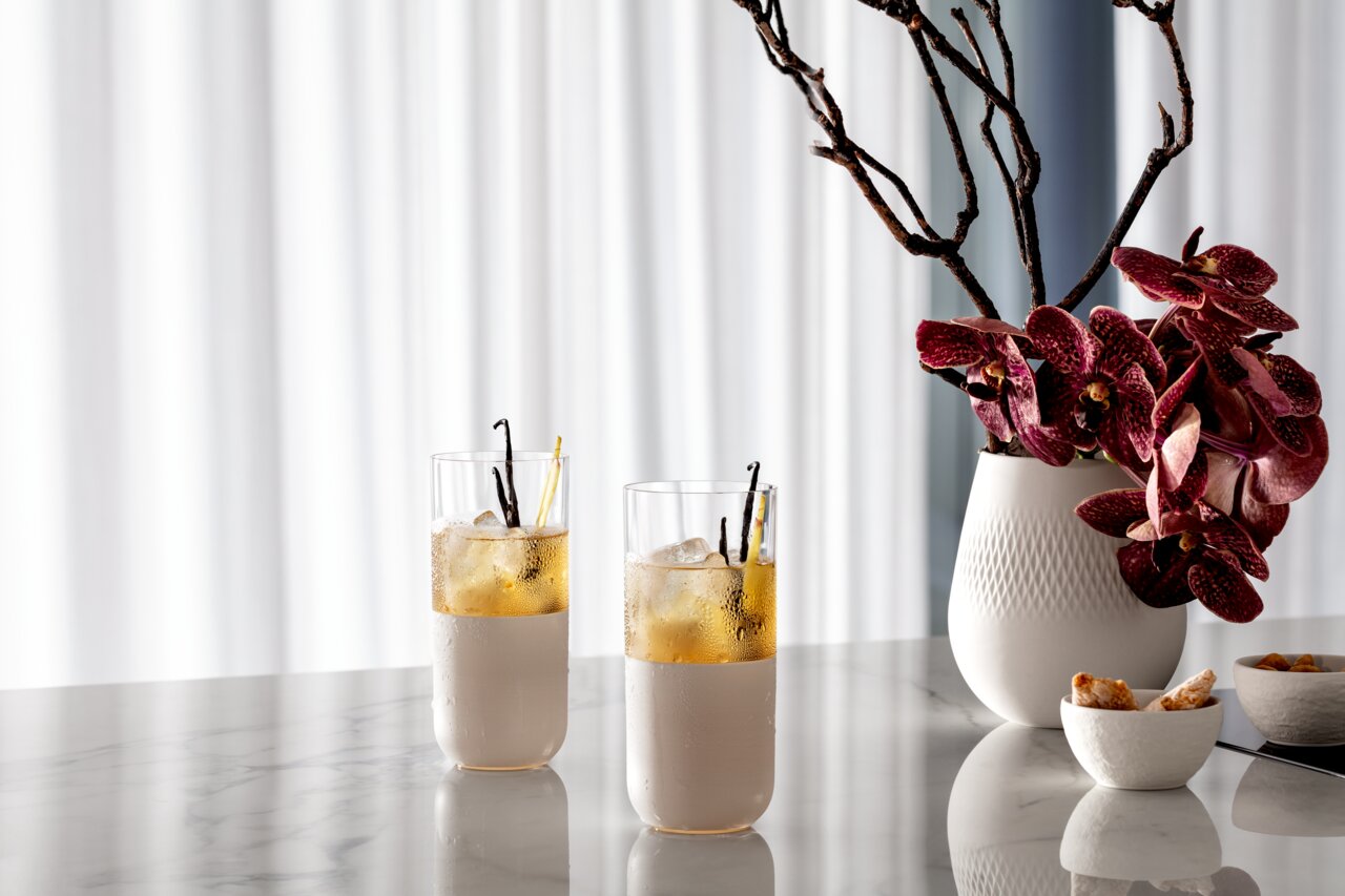Villeroy & Boch Longdrinkglas, 4 stuks, MANUFACTURE ROCK BLANC Twee longdrinkglazen met drankjes en ijsblokjes op een marmeren tafel, ernaast een vaas met paarse orchideeën, zijaanzicht.