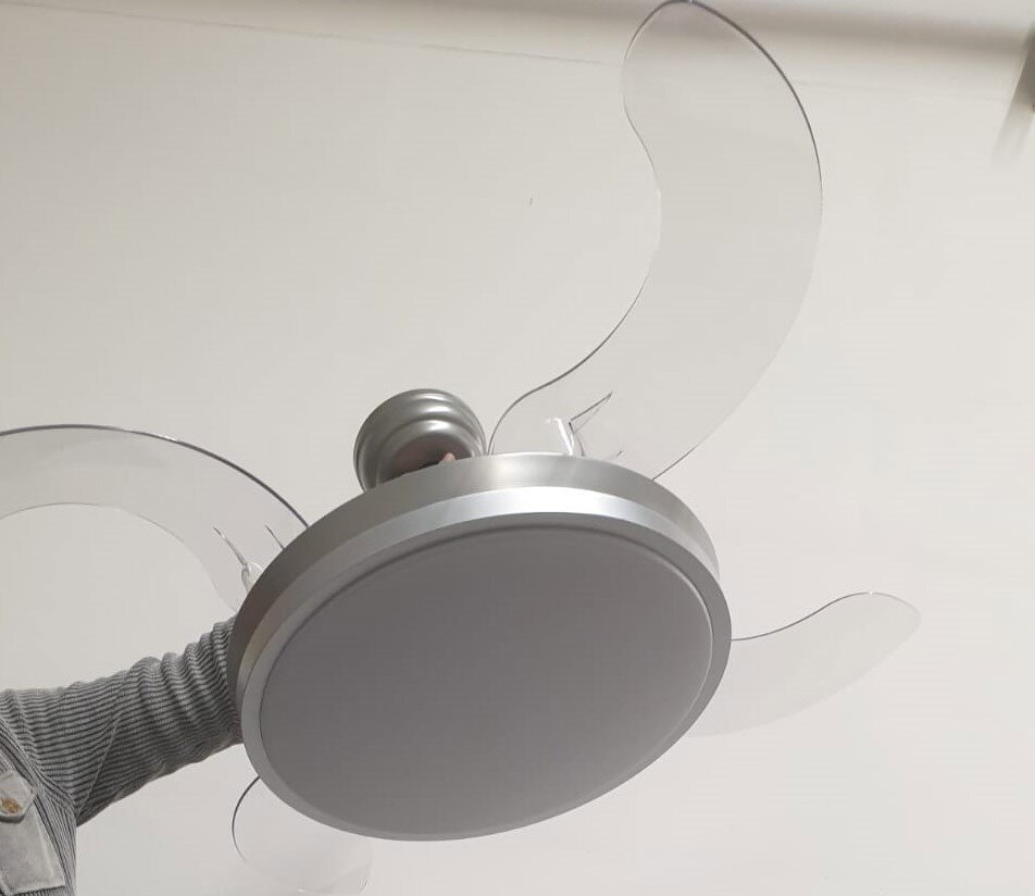 Moderner Deckenventilator mit integrierter runder LED-Leuchte und transparenten Flügeln in Silber/Weiß, von unten aus leichter Untersicht fotografiert.