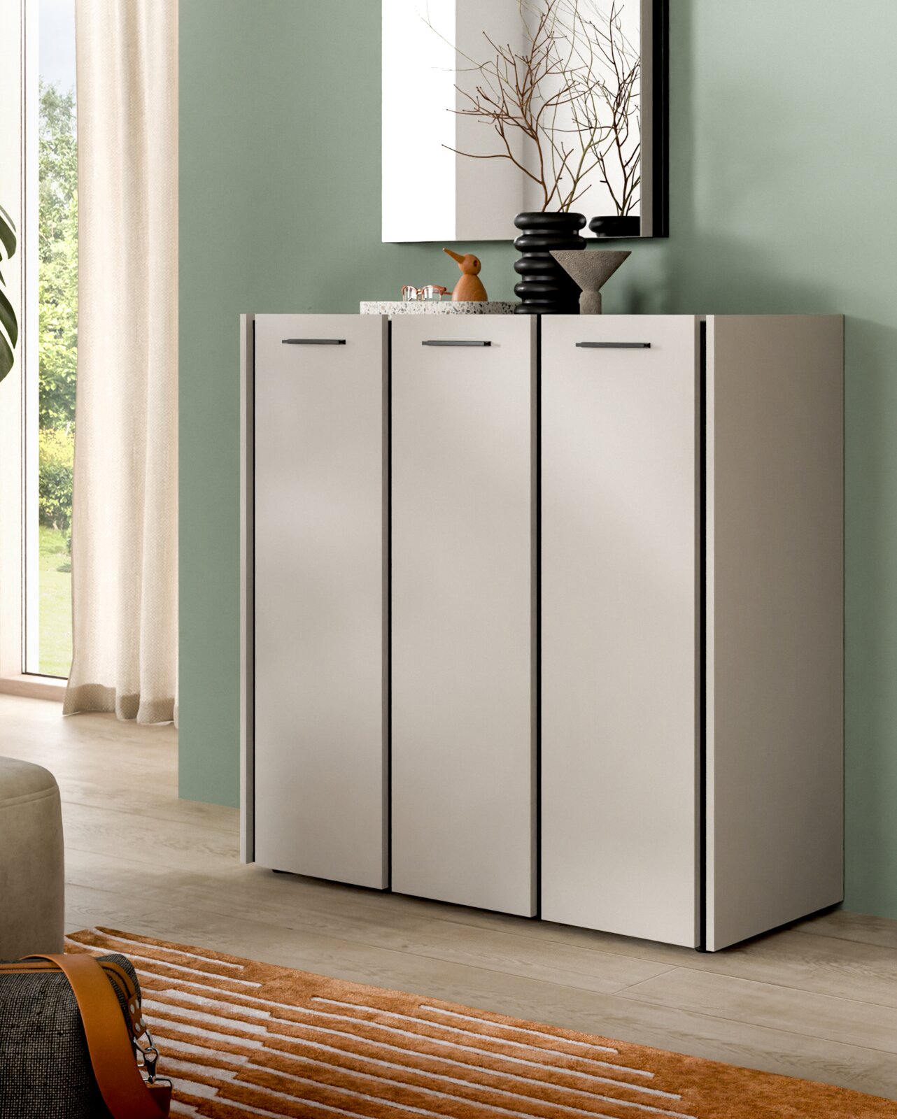 Modernes, weißes Sideboard mit drei Türen und schwarzen Griffen vor grüner Wand, dekoriert mit Vasen und Spiegel; Perspektive: frontale Ansicht in leichtem Winkel von links