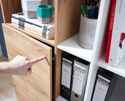 Nahaufnahme eines Aktenschranks mit Holz- und weißen Elementen, in dem Ordner und Bücher aufbewahrt werden. Eine Hand zeigt auf die Schranktür. Perspektive von schräg oben.