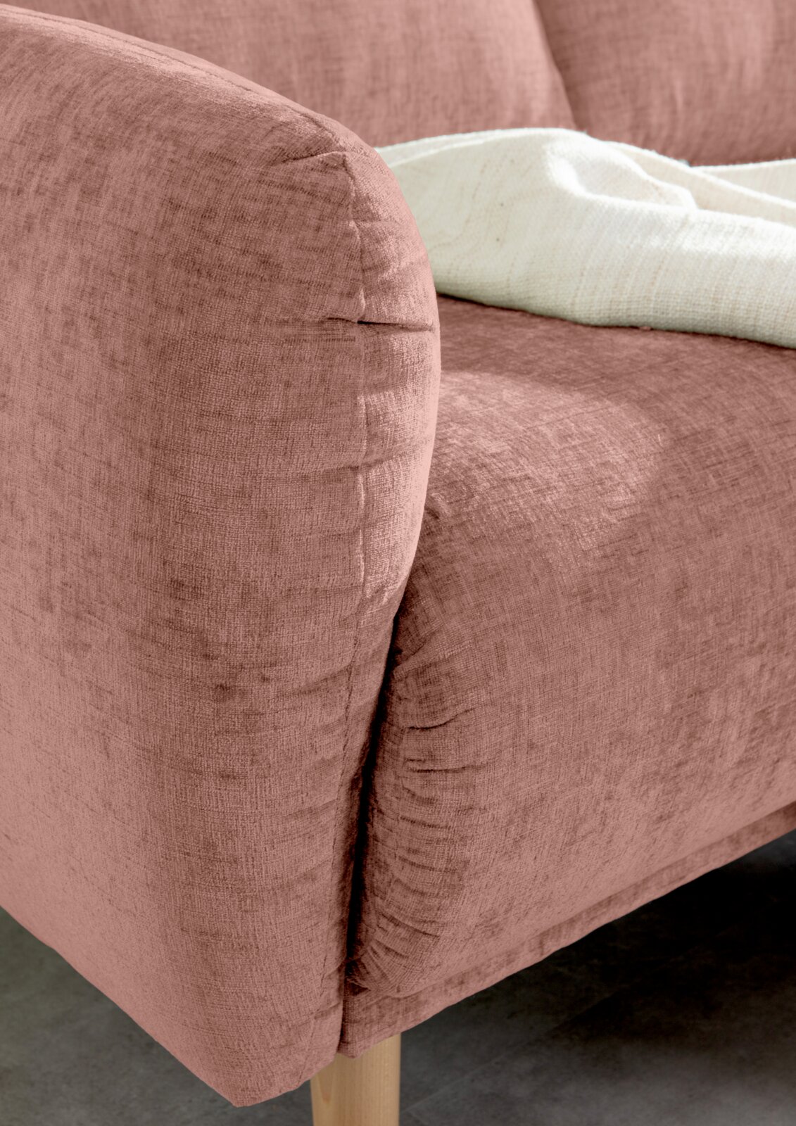 Nahaufnahme einer rosafarbenen Sofaecke mit weicher Textur und Holzfuß, von der Seite betrachtet.