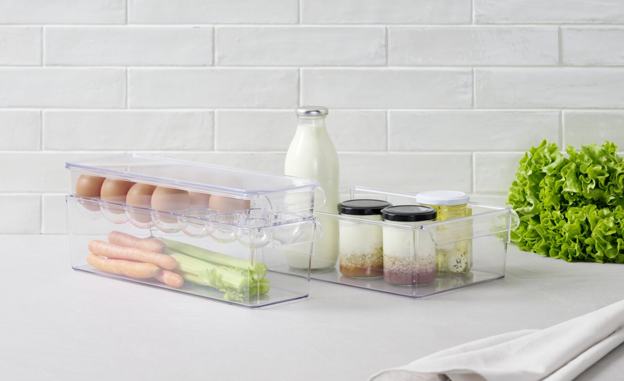 Transparenter 4-teiliger Organizer mit Eiern, Karotten und Sellerie in der Vorderansicht, daneben Gläser mit Joghurt und Milchflasche, auf einem Tisch mit Salat im Hintergrund.