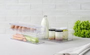 Transparenter 4-teiliger Organizer mit Eiern, Karotten und Sellerie in der Vorderansicht, daneben Gläser mit Joghurt und Milchflasche, auf einem Tisch mit Salat im Hintergrund.