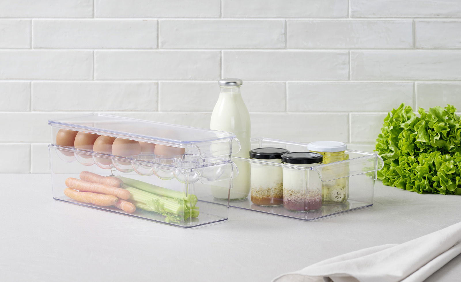 Transparenter 4-teiliger Organizer mit Eiern, Karotten und Sellerie in der Vorderansicht, daneben Gläser mit Joghurt und Milchflasche, auf einem Tisch mit Salat im Hintergrund.