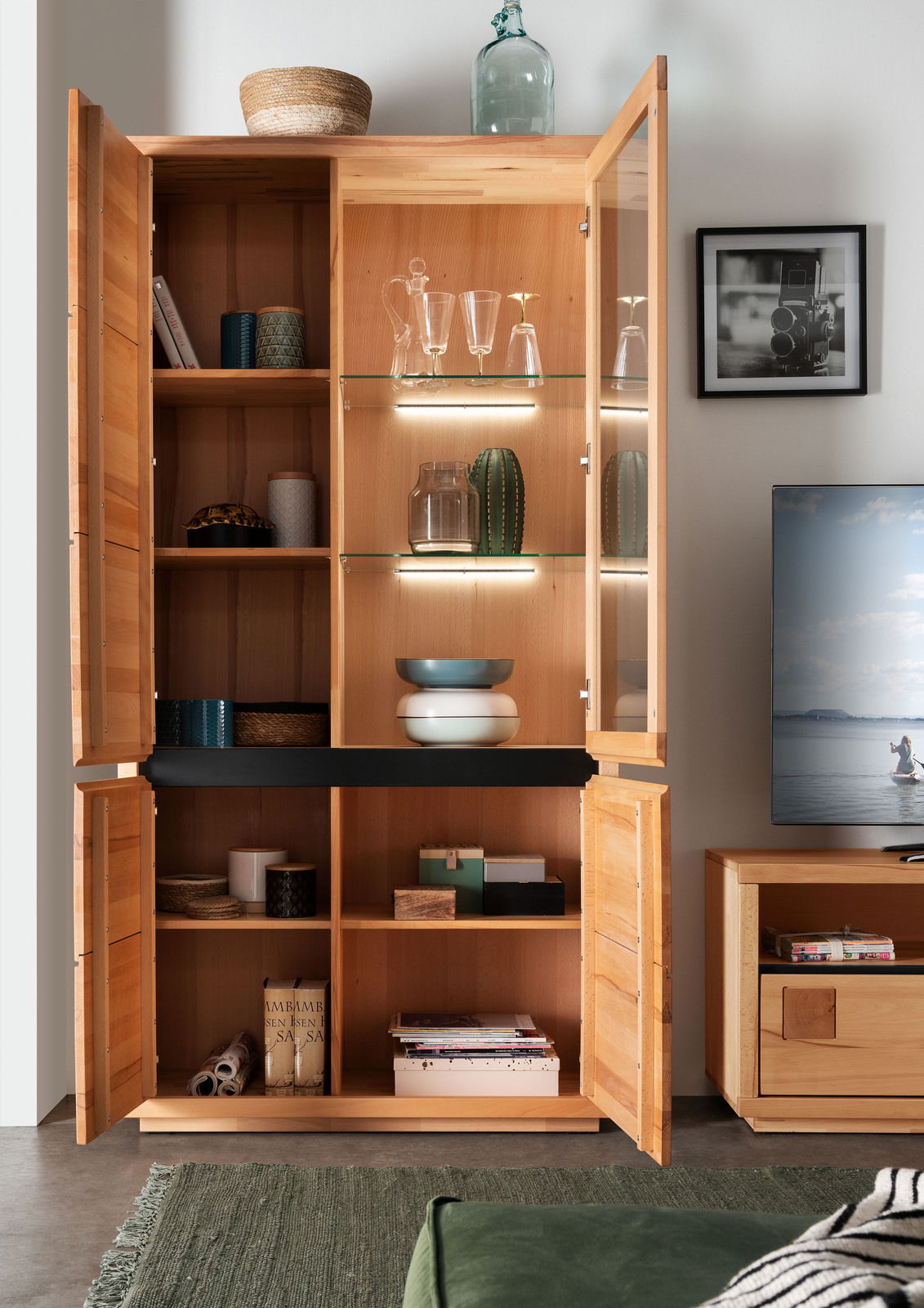 Geöffnete Vitrine aus Holz mit Glastüren, gefüllt mit Geschirr und Dekorationen, aus frontaler Perspektive.