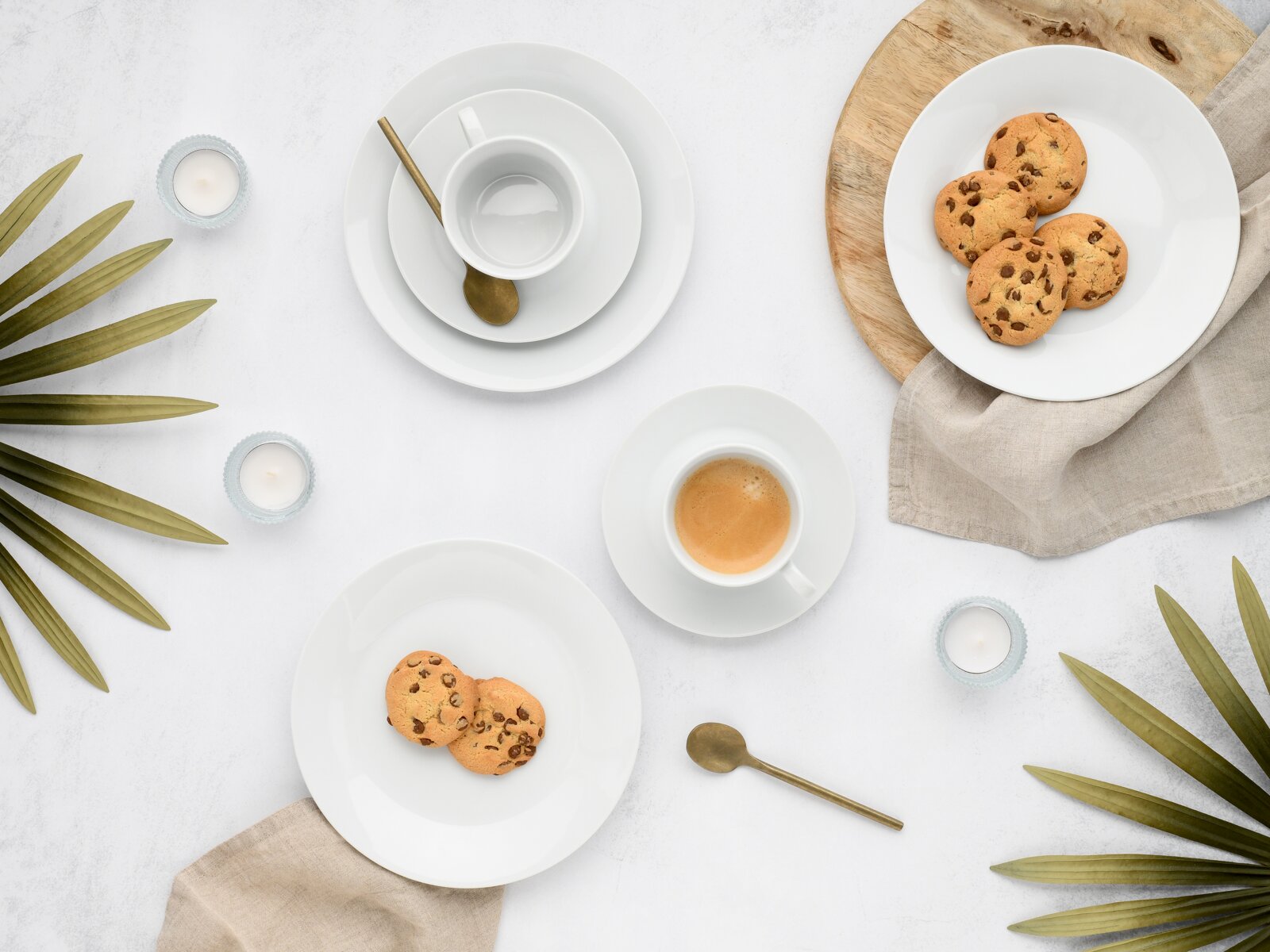 Minimalistisches Kaffeegedeck aus weißem Porzellan mit Espressotasse, Untertassen und Tellern, dazu goldene Löffel, Schokokekse und Teelichter auf hellem Hintergrund – Draufsicht (Flatlay)