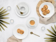 Minimalistisches Kaffeegedeck aus weißem Porzellan mit Espressotasse, Untertassen und Tellern, dazu goldene Löffel, Schokokekse und Teelichter auf hellem Hintergrund – Draufsicht (Flatlay)