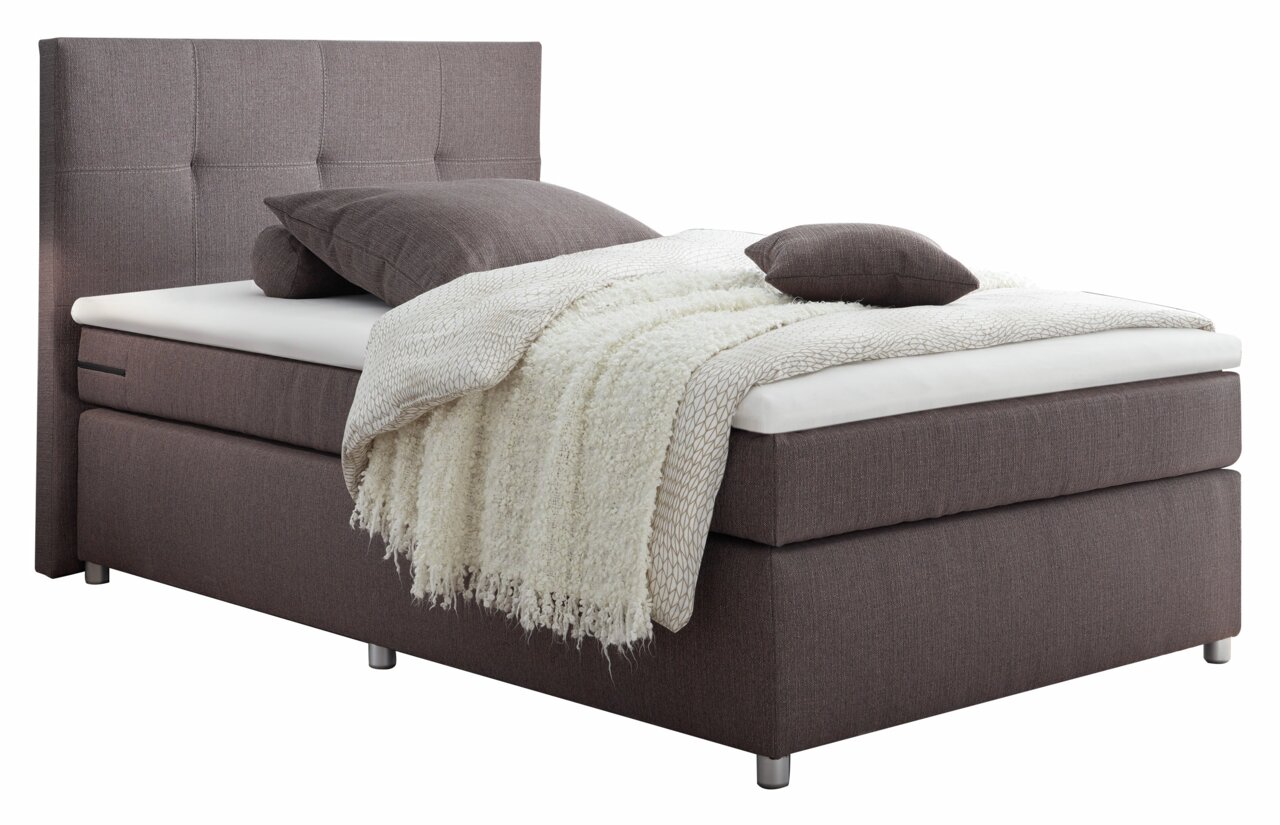 Boxspringbett in seitlicher Perspektive mit grauem Stoffbezug, Kopfteil mit quadratischer Steppung, ausgestattet mit Kissen und einer cremefarbenen Decke.