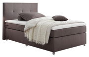 Boxspringbett in seitlicher Perspektive mit grauem Stoffbezug, Kopfteil mit quadratischer Steppung, ausgestattet mit Kissen und einer cremefarbenen Decke.