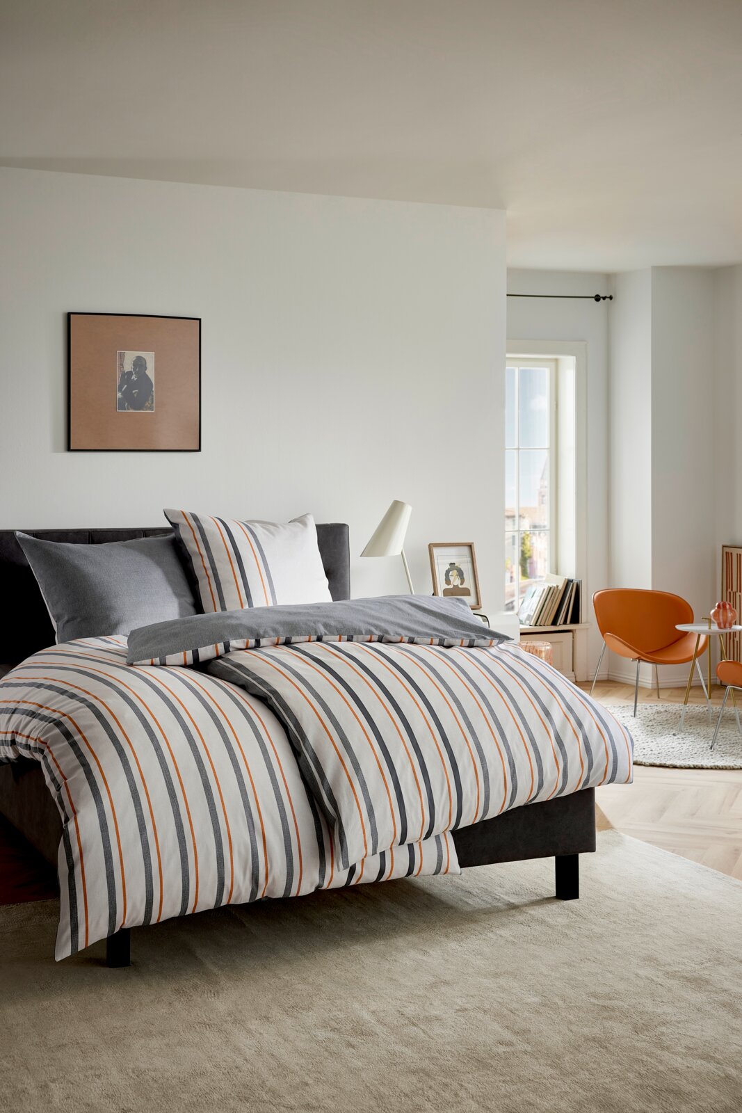 Gestreept dekbedovertrek in wit met grijze en oranje strepen op een donkergrijs bed in een lichte slaapkamer, gefotografeerd vanuit een schuin vooraanzicht.