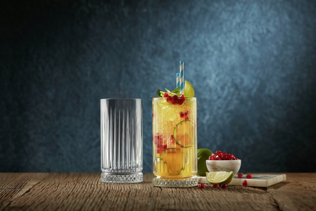 Zwei Longdrinkgläser Elysia auf einem Holztisch, eines leer und eines gefüllt mit einem erfrischenden Getränk, garniert mit Limetten, Granatapfelkernen und einem blauen Strohhalm, seitliche Perspektive.