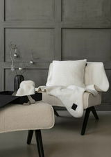 Gemütliches Geschenkset mit weißem Kissen und Decke auf einem Sessel, mit schwarzem Geschenkkarton im Vordergrund, seitliche Perspektive.