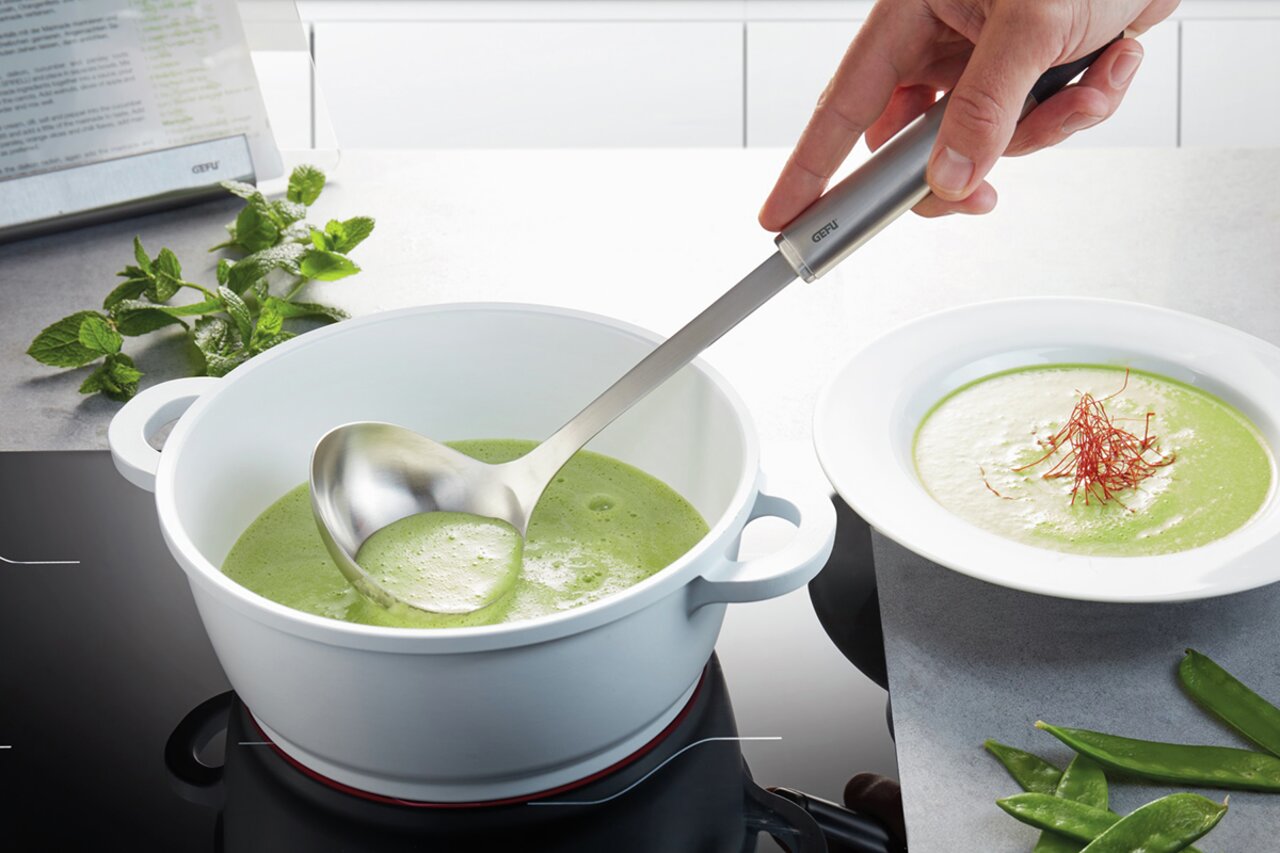 Hand hält einen Schöpflöffel PRIMELINE über einem Topf mit grüner Suppe, seitliche Perspektive