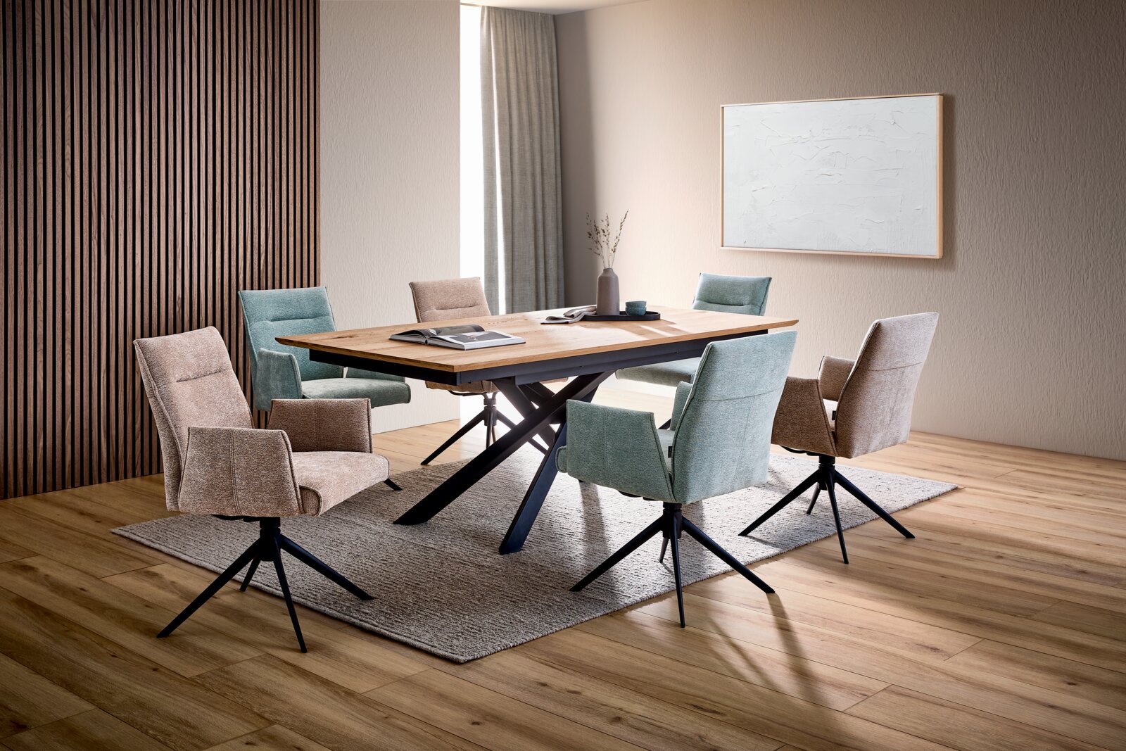 Modernes Esszimmer-Set mit großem Esstisch aus Holz und schwarzem X-Gestell, umgeben von gepolsterten Drehstühlen in Mintgrün und Beige; Perspektive: schräg von vorne auf Tisch und Stühle.