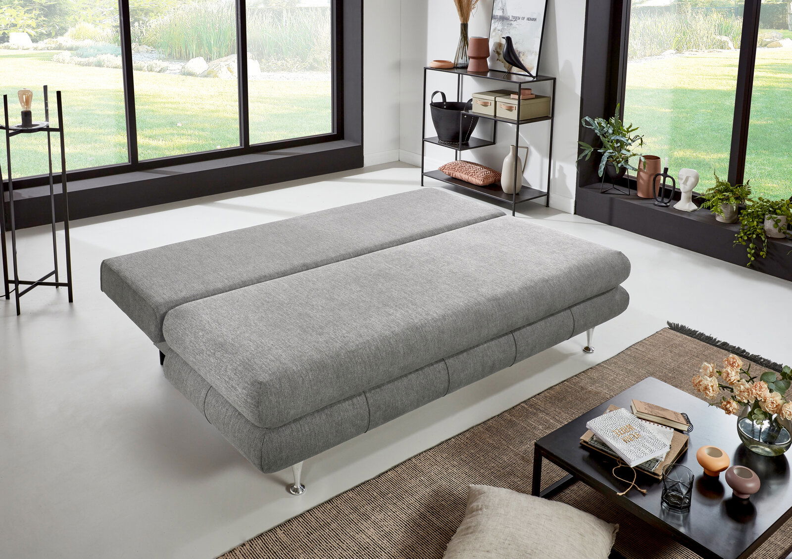 Graues Funktionssofa in flacher Position, seitliche Perspektive, in modernem Wohnzimmer mit großen Fenstern und stilvoller Dekoration.