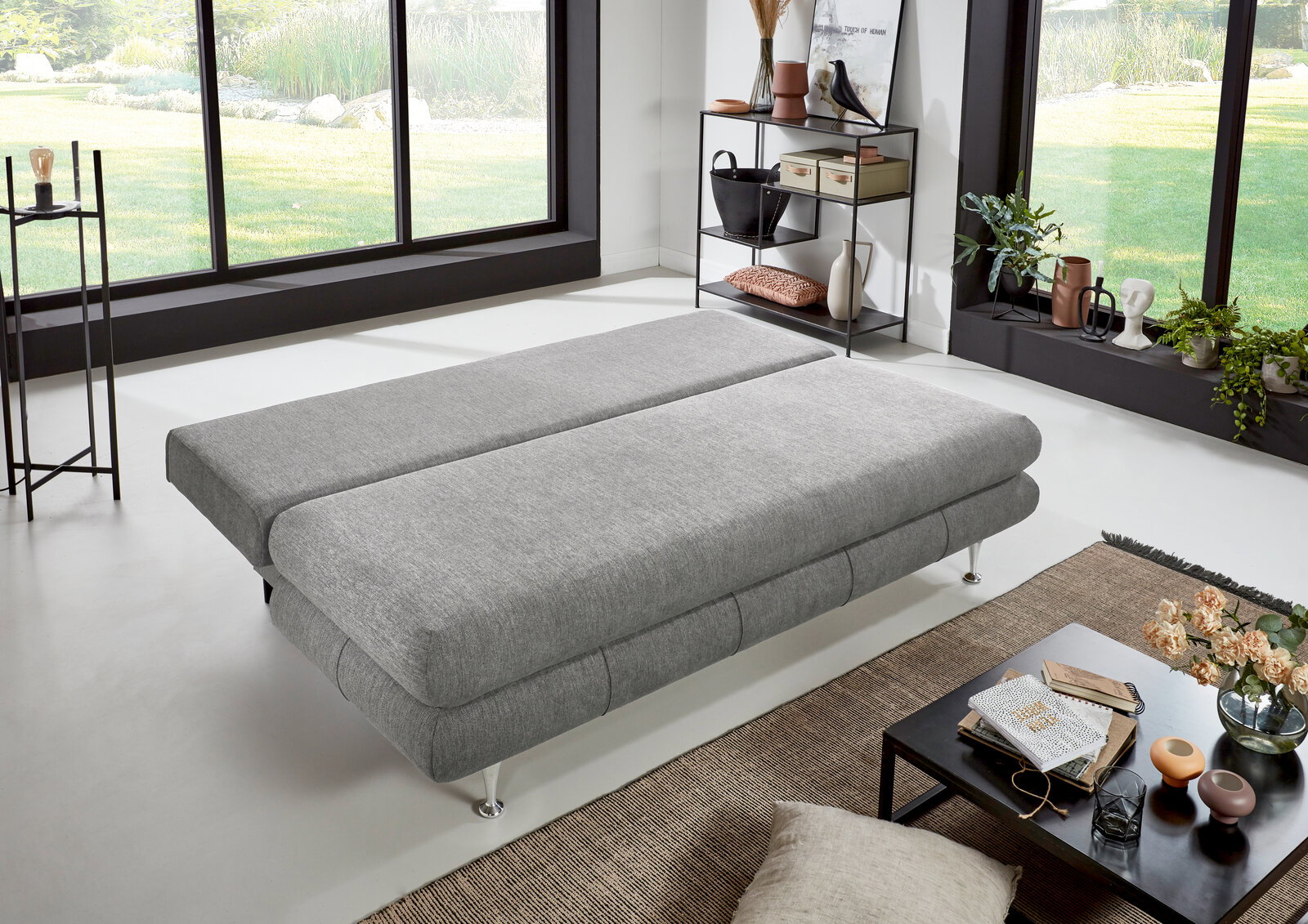 Graues Funktionssofa in flacher Position, seitliche Perspektive, in modernem Wohnzimmer mit großen Fenstern und stilvoller Dekoration.