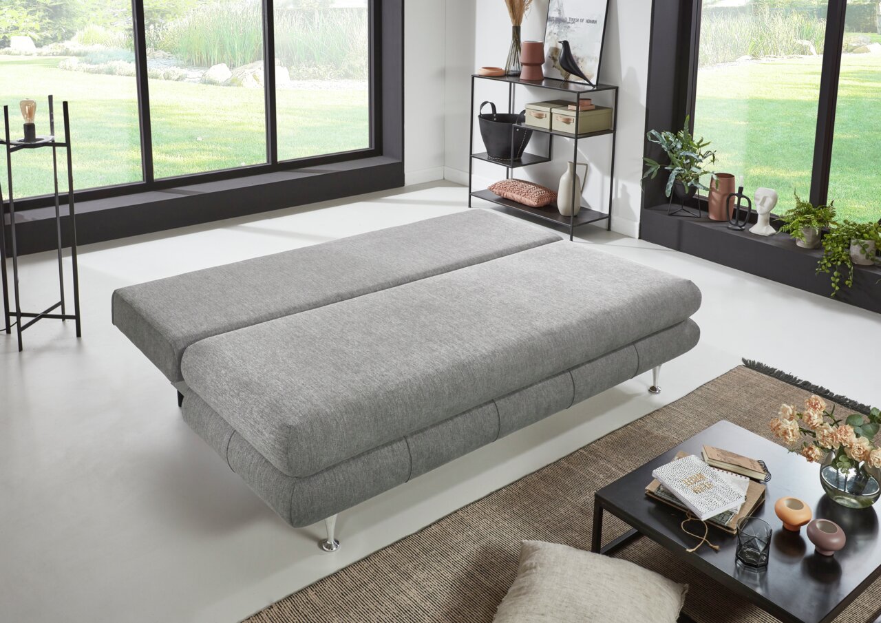 Graues Funktionssofa in flacher Position, seitliche Perspektive, in modernem Wohnzimmer mit großen Fenstern und stilvoller Dekoration.