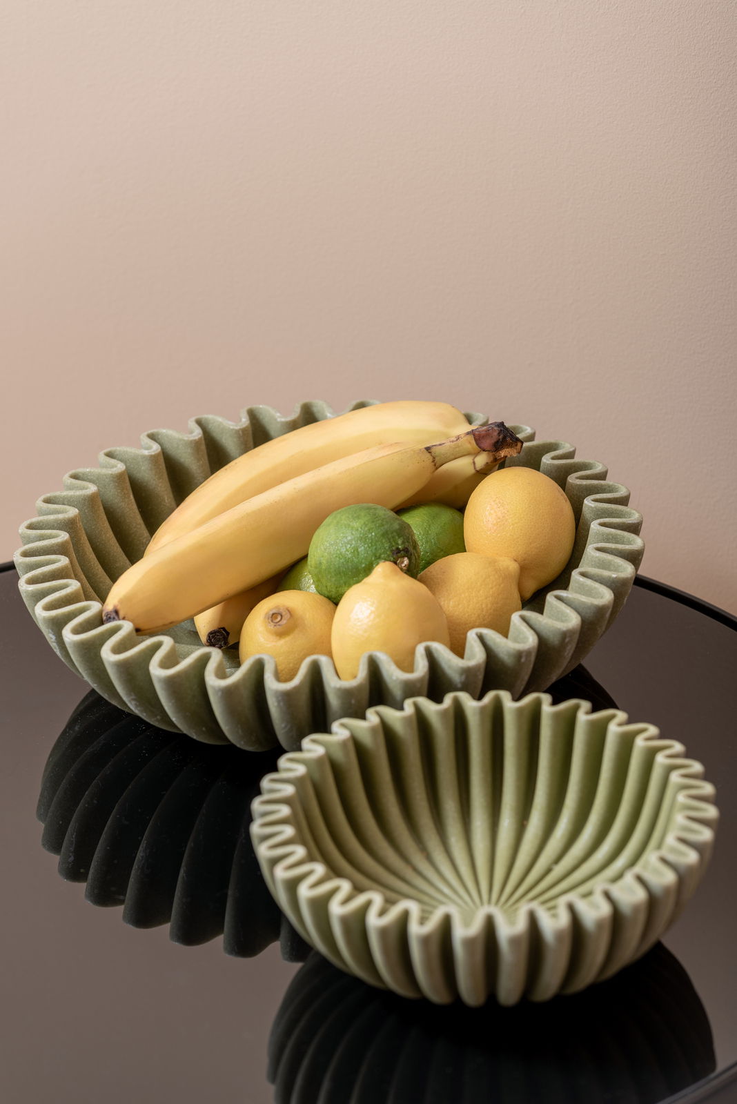 Zwei grüne Schalen mit gewelltem Rand, eine gefüllt mit Bananen, Zitronen und Limetten, auf einem glänzenden schwarzen Tisch aus der Vogelperspektive.
