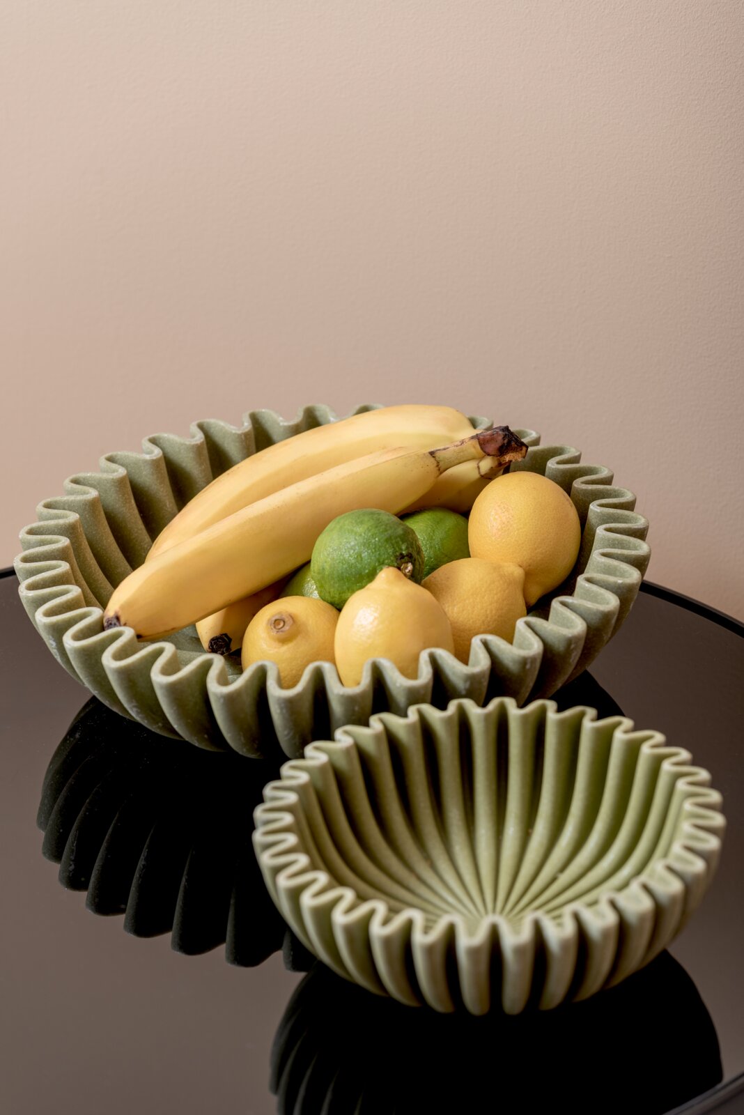 Zwei grüne Schalen mit gewelltem Rand, eine gefüllt mit Bananen, Zitronen und Limetten, auf einem glänzenden schwarzen Tisch aus der Vogelperspektive.