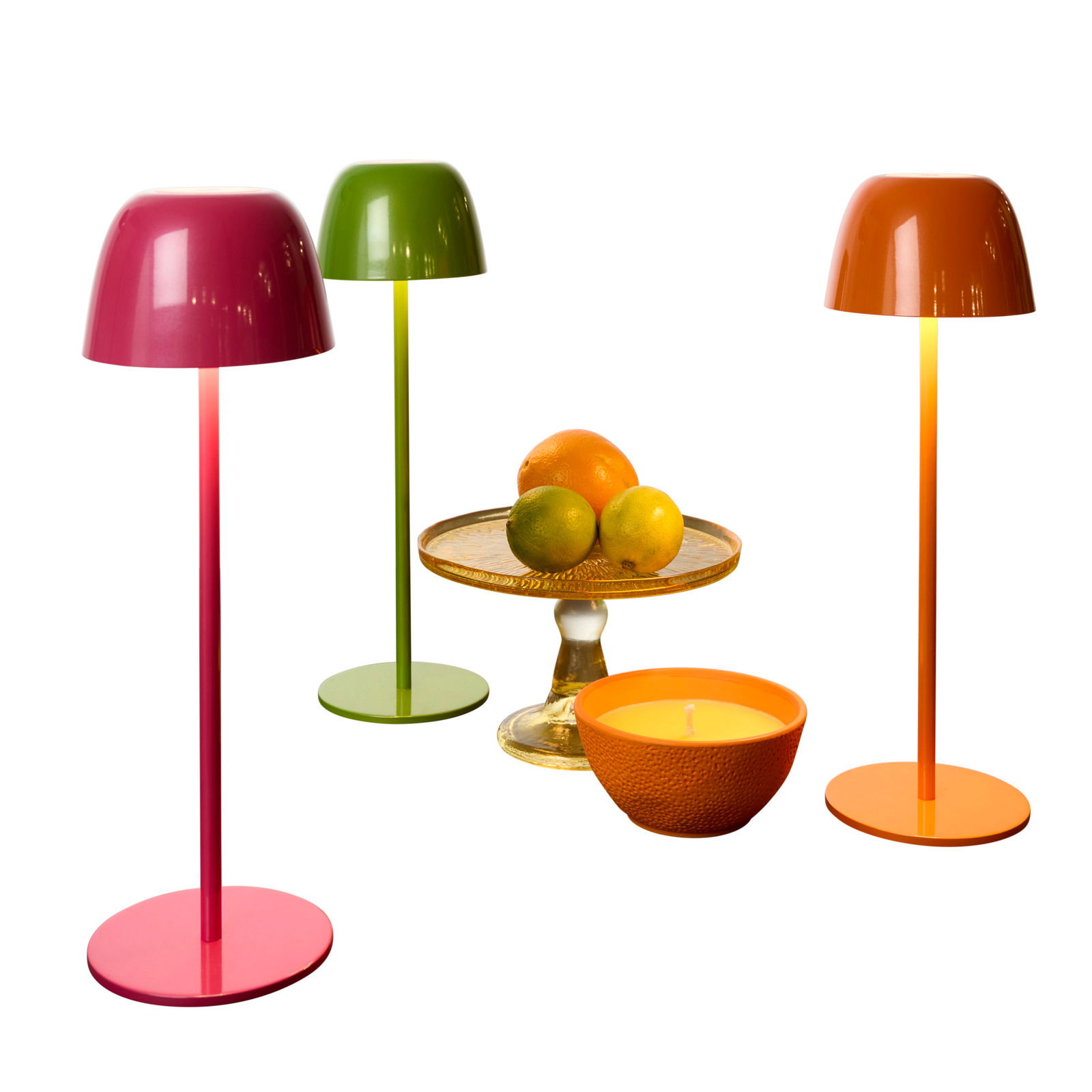 Novel Akku-Tischleuchte SANTA ANA Drei bunte Akku-Tischleuchten in Rot, Grün und Orange, frontal betrachtet, neben einer Obstschale und einer Kerze.