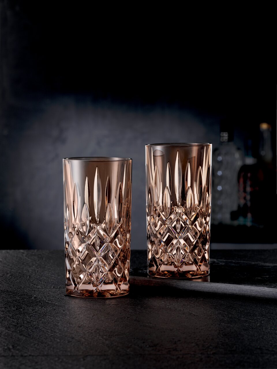 Twee elegante longdrinkglazen in het Noblesse-ontwerp, van kristalglas met een gefacetteerd patroon, op een donkere ondergrond, zijaanzicht.