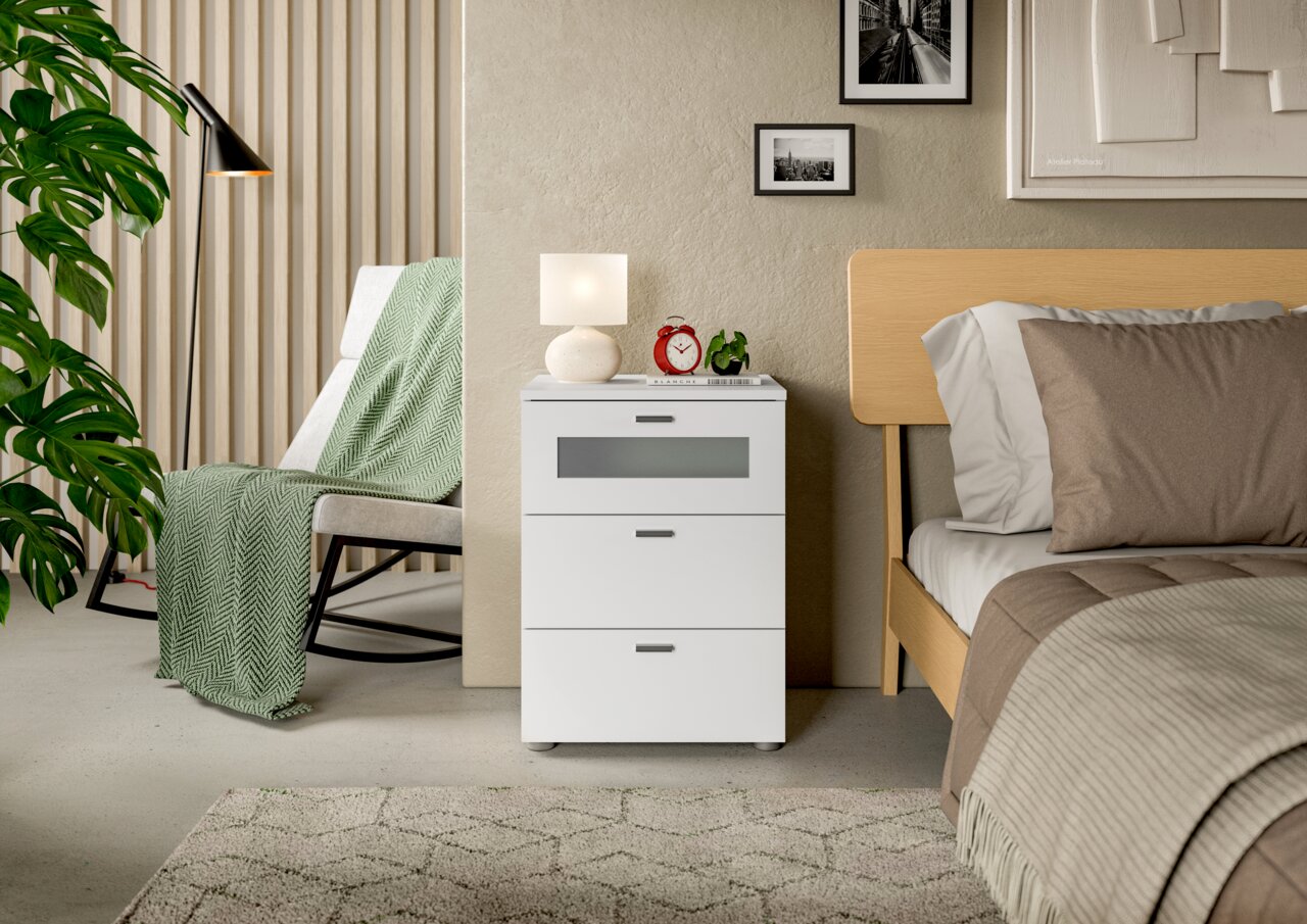 Weiße Nachtkommode mit drei Schubladen, dekoriert mit einer Lampe und einem Wecker, neben einem Bett in einem modernen Schlafzimmer, seitliche Perspektive.