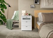 Weiße Nachtkommode mit drei Schubladen, dekoriert mit einer Lampe und einem Wecker, neben einem Bett in einem modernen Schlafzimmer, seitliche Perspektive.
